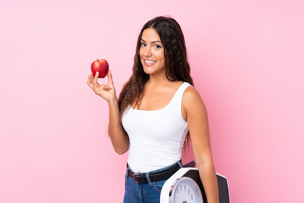 Young woman over isolated pink  with weighing machine and with an apple