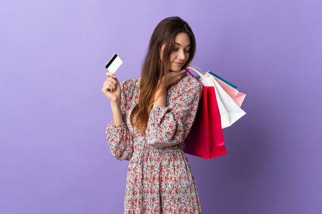 Young woman isolated on purple wall holding shopping bags and a credit card