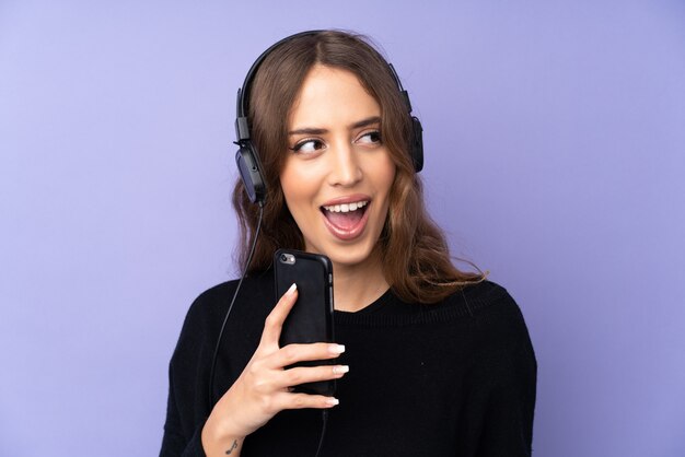 携帯電話と歌で音楽を聴く分離の紫色の壁の上の若い女性