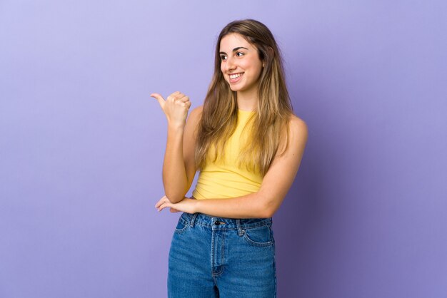 Young woman over isolated purple wall pointing to the side to present a product