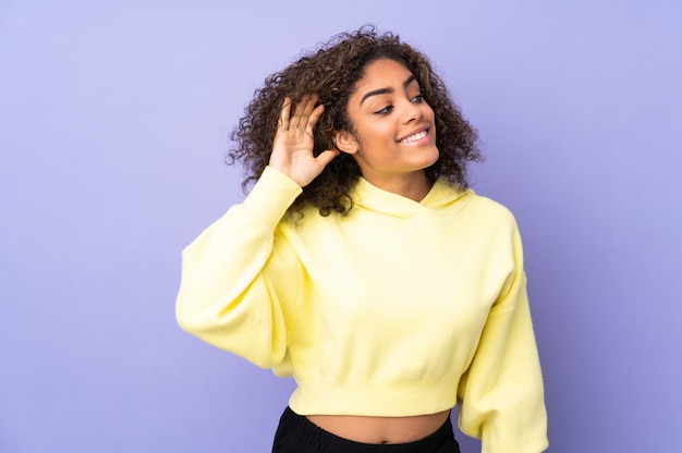 Young woman isolated on wall listening to something by putting hand on the ear