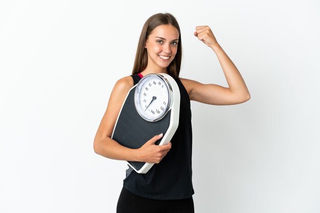 Young woman over isolated white background holding a weighing machine and doing strong gesture