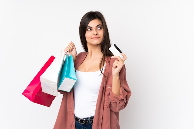 Young woman over isolated white holding shopping bags and a credit card and thinking