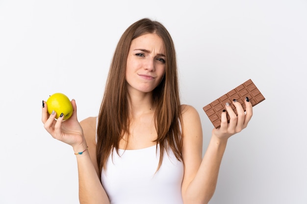 Молодая женщина над изолированной белой стеной, сомневаясь, принимая шоколадную таблетку в одной руке и яблоко в другой