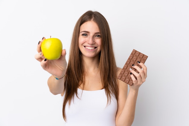 Young woman over isolated white wall taking a chocolate tablet in one hand and an apple in the other