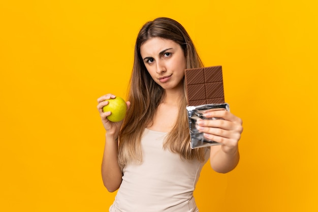 Молодая женщина на изолированном желтом, принимая шоколадную таблетку в одной руке и яблоко в другой