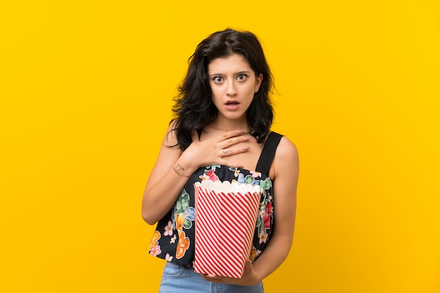 Young woman over isolated yellow wall holding a bowl of popcorns