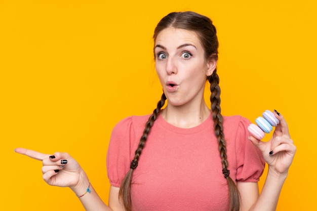 Young woman over isolated yellow wall holding colorful French macarons and pointing side