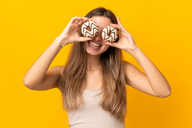 Young woman over isolated yellow wall holding donuts in eyes