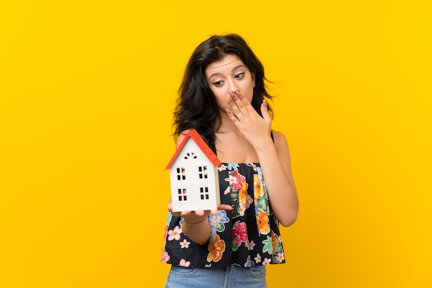 Young woman over isolated yellow wall holding a little house