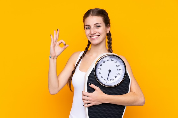 Young woman over isolated yellow wall holding a weighing machine and doing OK sign