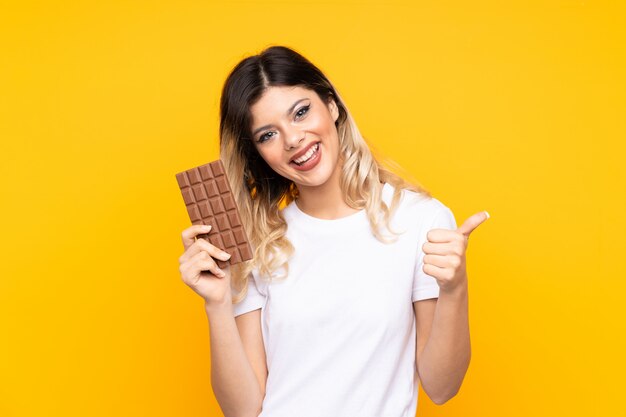 Молодая женщина, изолированные на желтой стене, принимая шоколадные таблетки и с пальца вверх