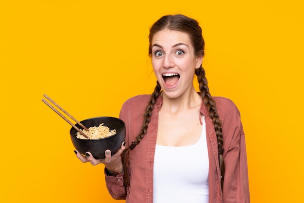 Young woman over isolated yellow wall with surprise and shocked facial expression while holding a bowl of noodles with chopsticks