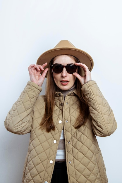 Молодая женщина в куртке в шляпе в солнечных очках на фоне белой стены