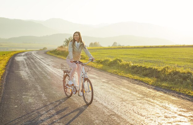 ジャケットを着た若い女性が田舎道を自転車に乗り、午後の日差しが背景に輝く