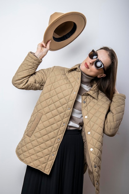 A young woman in a jacket takes off her hat near a white\
wall