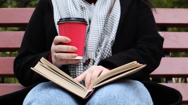 Young woman in jeans, coat and scarf, on a park bench. A woman is reading a book and drinking coffee or other hot drink outdoors alone. Close-up. The concept of honor, study, leisure and recreation.