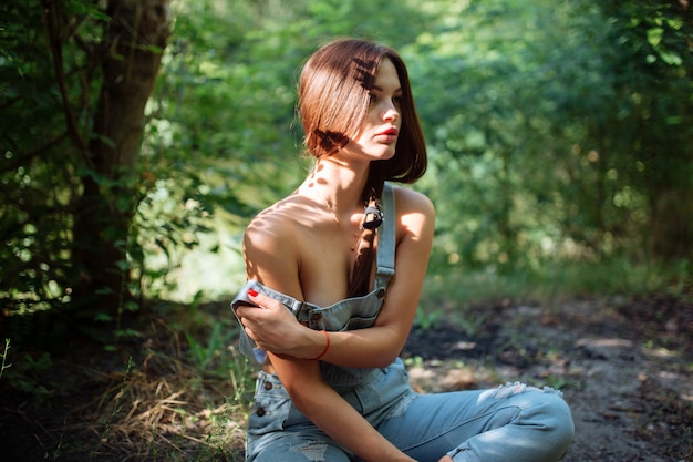 Young woman in jeans overalls standing under tree in woodland.