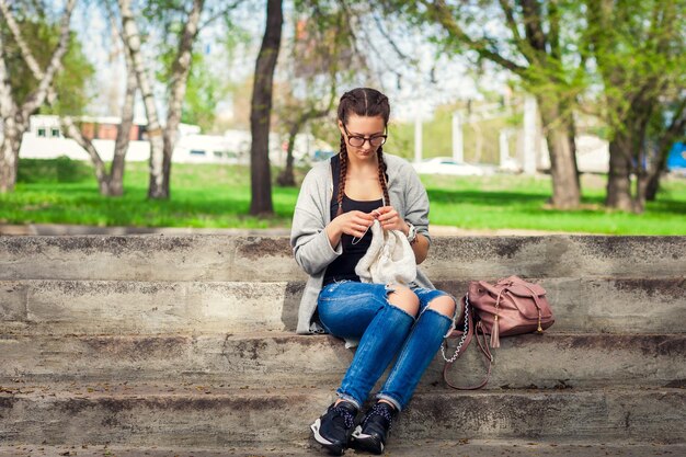young woman in jeans and with pigtails knits a sweater in the city on the grass