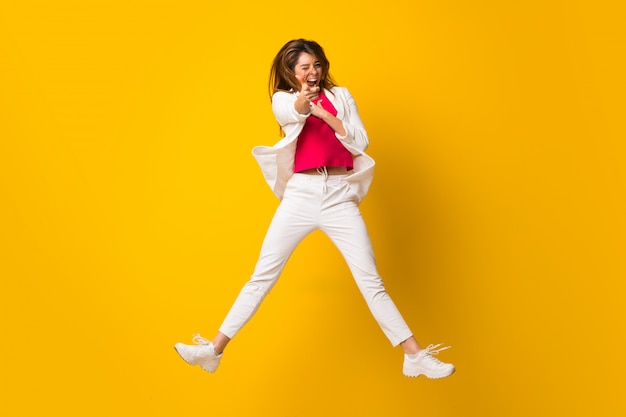 Young woman jumping over isolated yellow wall pointing to the front