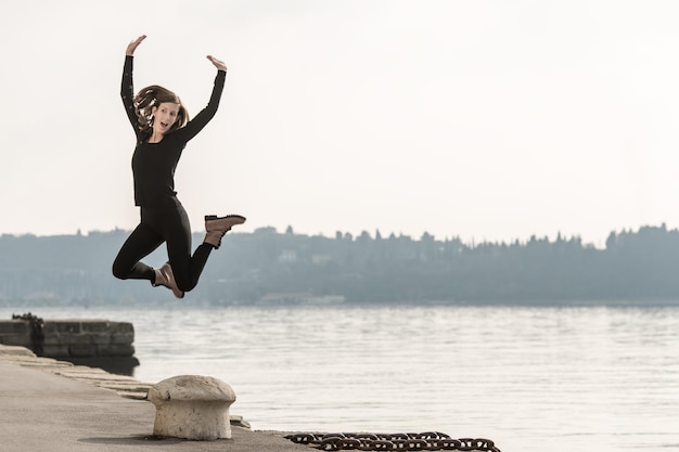 Young woman jumping for joy above a bollard on a quay overlooking a lake or the ocean on a misty atmospheric day