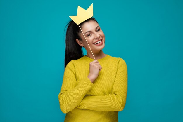 Young woman keeping paper crown and posing on blue isolated background. 