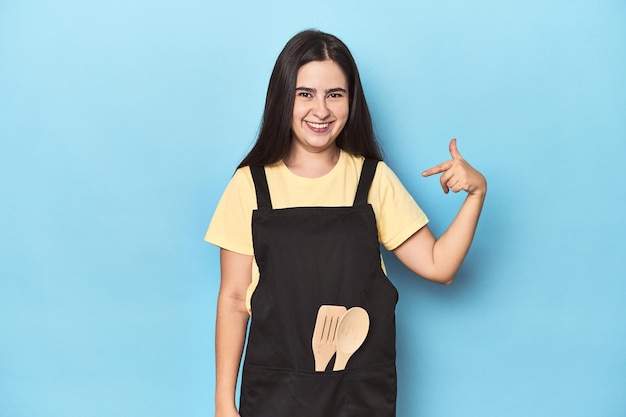 Young woman in kitchen apron on blue person pointing by hand to a shirt copy space proud