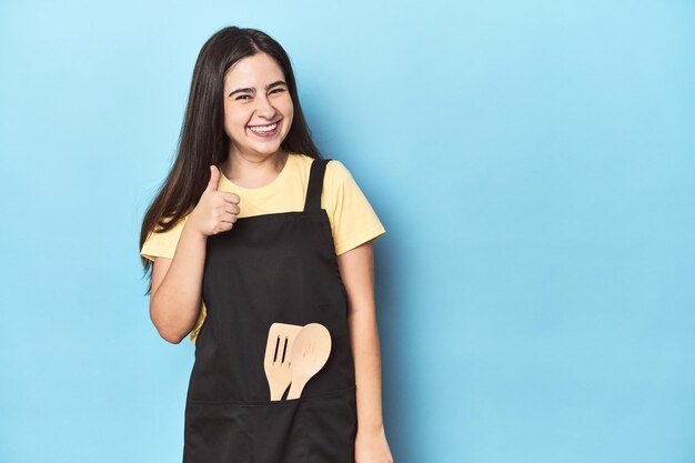 Photo young woman in kitchen apron on blue smiling and raising thumb up