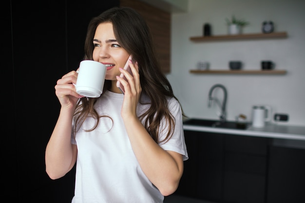 Young woman in kitchen during qiarantine. Talking on phone and drink from white cup tea or coffee. Online conversation wireless. Modern technologies and devices.