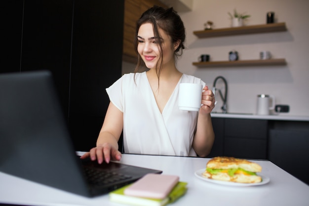 Young woman in kitchen during quarantine. Girl sit at table and using laptop. Hold white cup in hand. Working remote from home office.