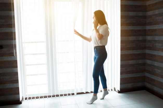 Young woman on kitchen during quarantine. Stand at window on tip of her toes and look outside through curtains. Talking on phone. Alone in room.