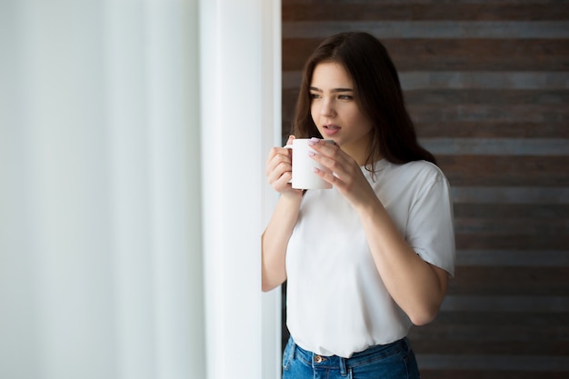 Giovane donna in cucina. stare alla finestra e bere caffè o tè. guarda fuori attraverso la tenda. calma pacifica donna sola nella stanza.