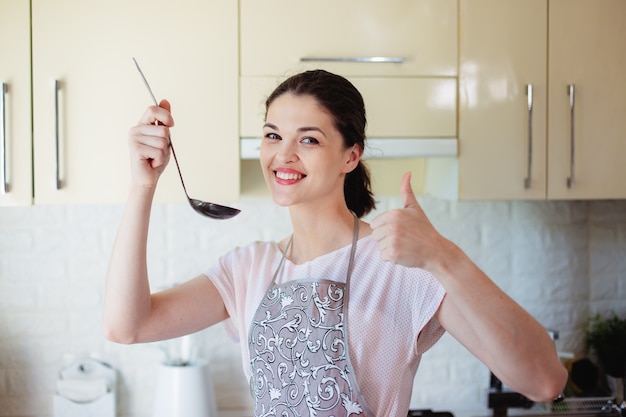 Young woman in the kitchen tastes soup on salt with a ladle. showing thumb