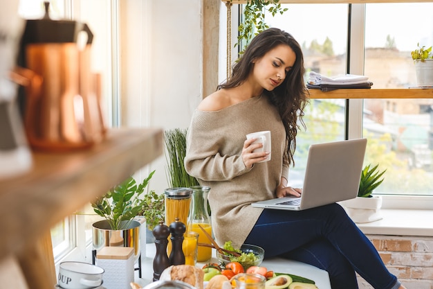 ラップトップコンピューターのレシピを探して、笑顔のキッチンで若い女性。フードブロガーのコンセプト