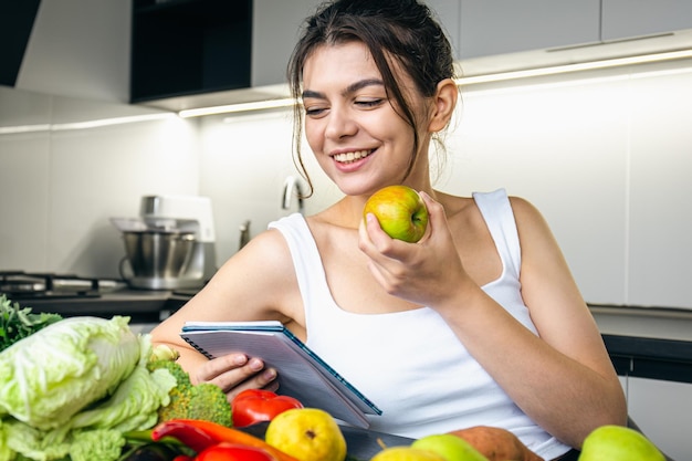 A young woman in the kitchen with a notebook in her hands eats an apple