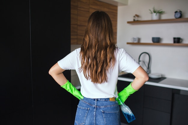 Young woman in kitchene. Back view ob brunette wear green protect gloves for cleaning. Hold cloth in hand. Ready to clean kitchen.