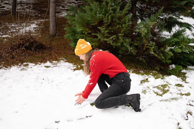 A young woman in knitted clothes collects snowballs for a snowball game against a background of green Christmas trees outdoors The concept of having fun and winter activities in nature