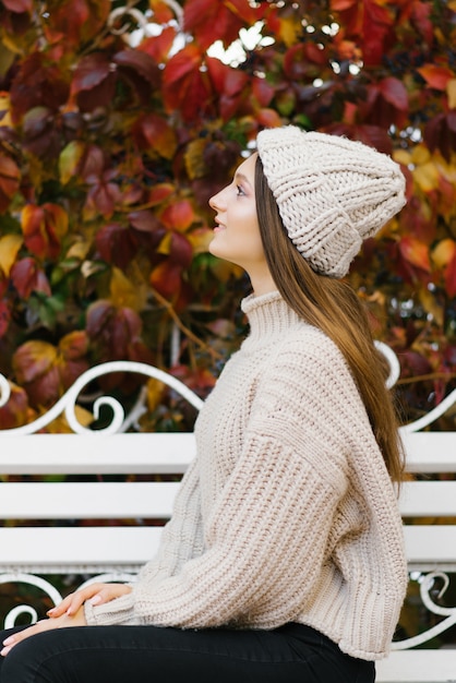 A young woman in a knitted sweater and hat sits on a white bench in an autumn Park and dreams