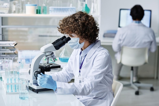 Young woman in labcoat protective mask and gloves looking in microscope