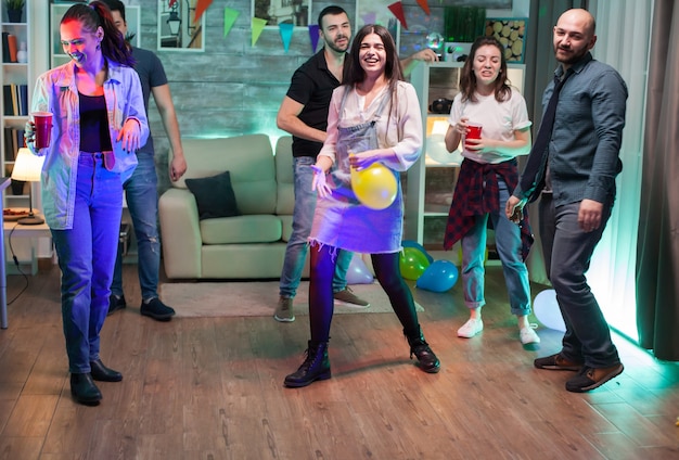Молодая женщина смеется из-за воздушного шара гелия, танцуя на вечеринке со своими друзьями.