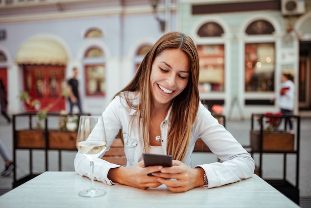 Young woman laughing while using phone at outdoor restaurant.