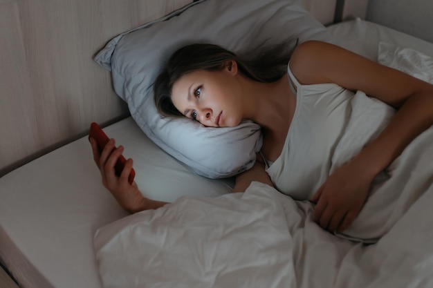 写真 ベッドに横になって夜にスマートフォンを見ている若い女性