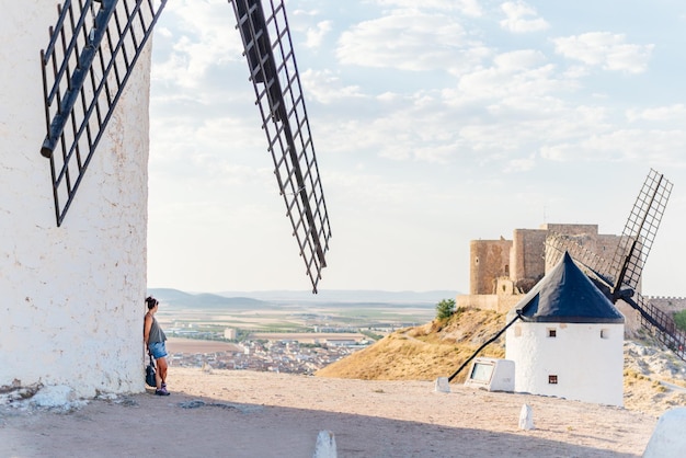 Young woman leaning against the facade of a windmill in the\
shade while contemplating the panorama with windmills and a\
medieval castle in toledo, spain.