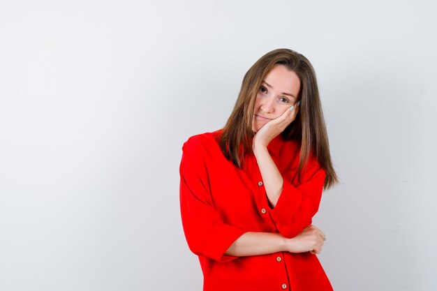 Young woman leaning cheek on hand in red blouse and looking thoughtful. front view.