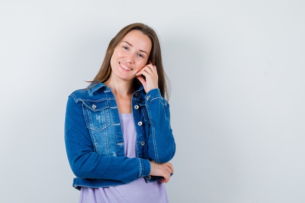 Tシャツ、ジャケット、陽気に見える手に頬をもたれている若い女性。正面図。