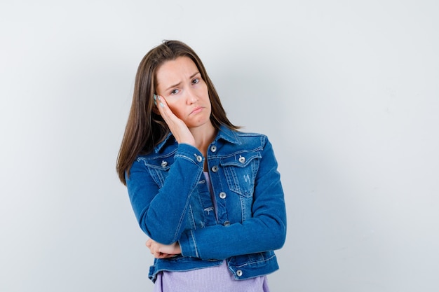 Young woman leaning cheek on hand in t-shirt, jacket and looking disappointed , front view.