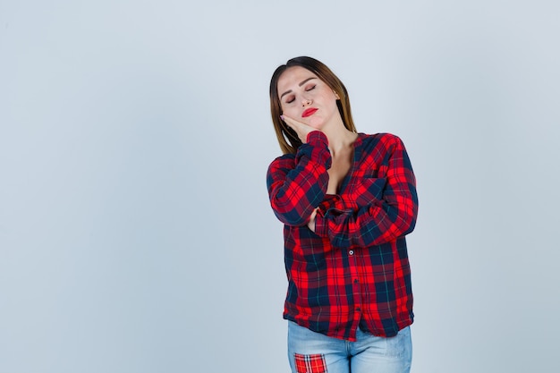 Giovane donna appoggiata guancia sul palmo in camicia a quadri, jeans e guardando allegro. vista frontale.