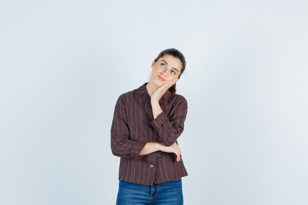 Giovane donna appoggiata guancia sul palmo, pensando a qualcosa in camicia a righe, jeans e guardando pensieroso, vista frontale.