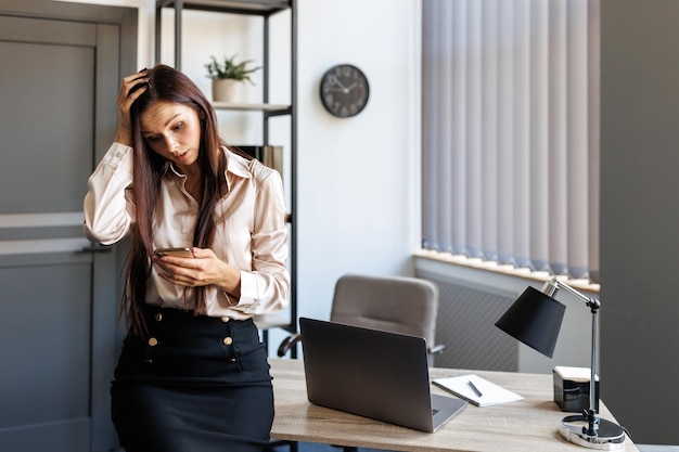 Young woman leaning on a desk holding a mobile phone and with a sad look on her face looking at the screen reading and writing a message and holding her head with her hand