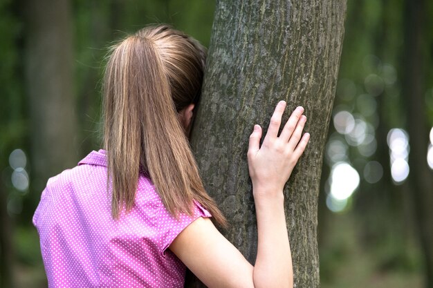 Young woman leaning to tree trunk hugging it with her hands in summer forest.